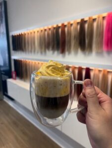 Hand holding a specialty coffee in front of a wall of high-quality hair extensions at Fusion Salon.